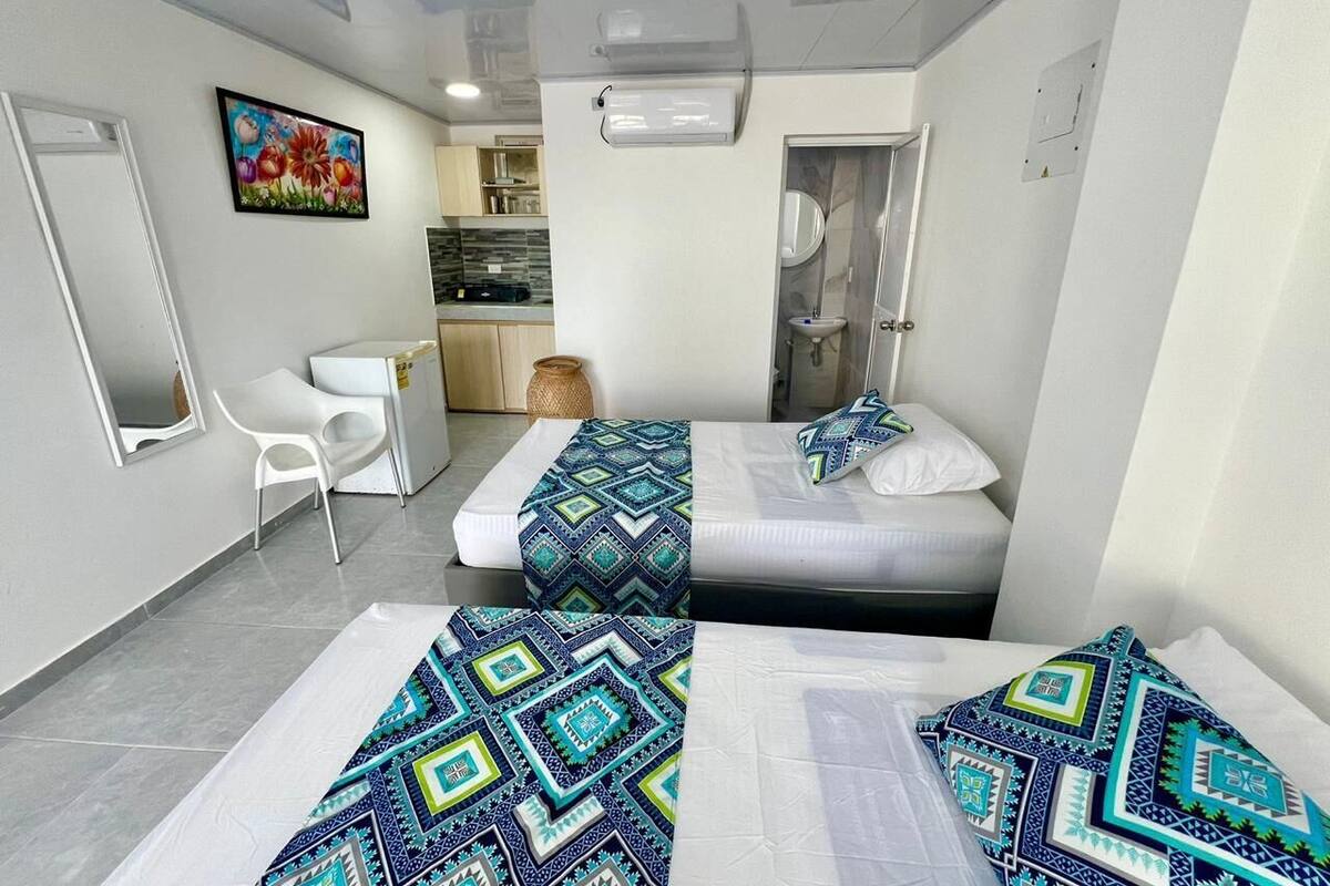 A hotel room with two beds, a small kitchenette, a white chair, and a painting on the wall.