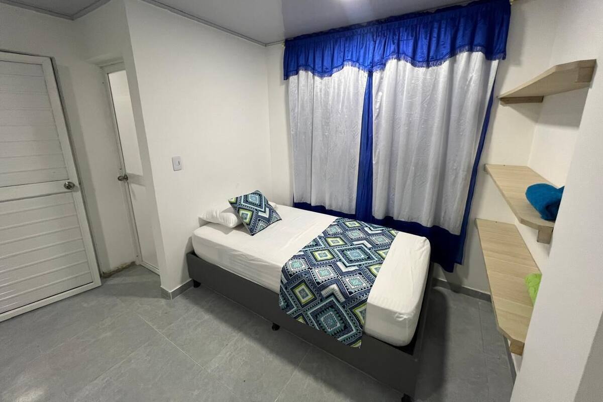 A small, clean room with a bed, a window with blue curtains, a wooden shelf, and a door.