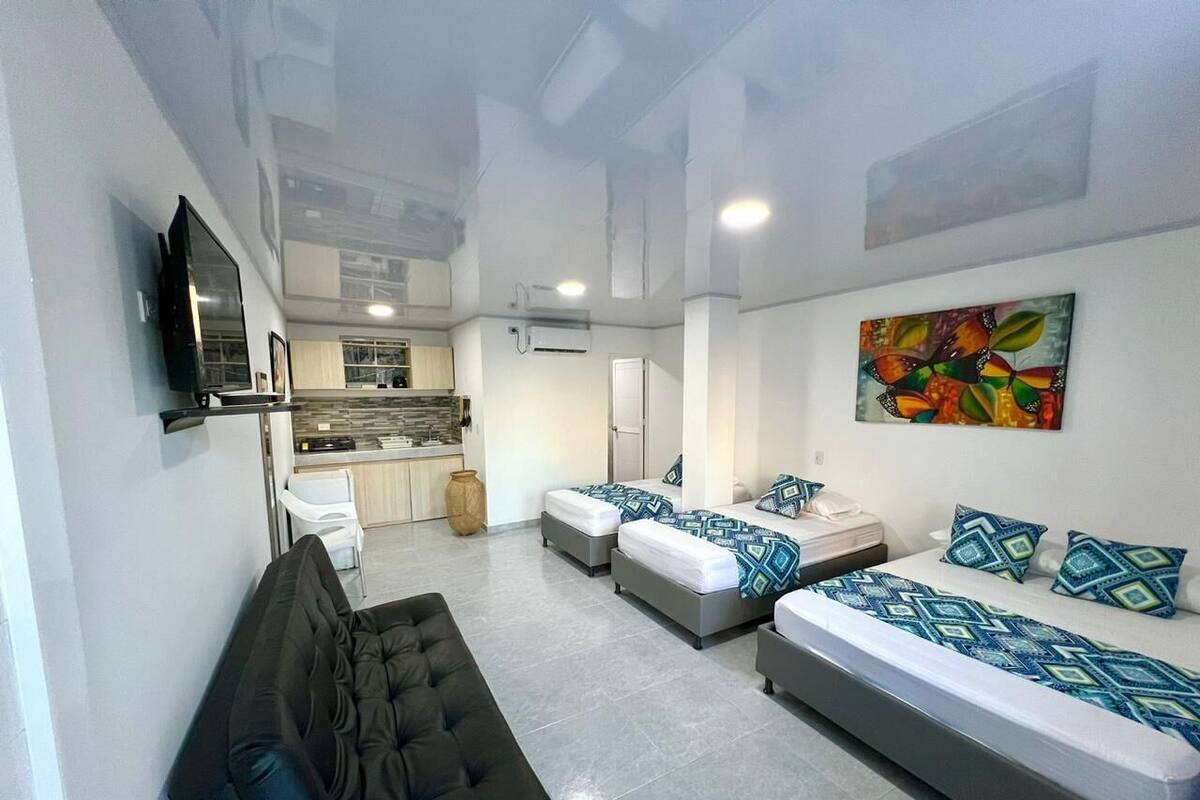 A modern hotel room with a sofa, two beds with patterned bedding, a kitchen area, and a large window.