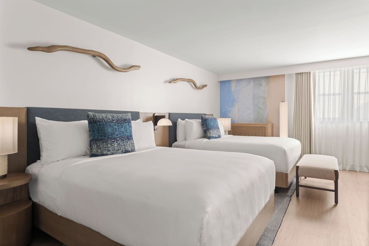 A hotel room with two beds, a small stool, and a large window with curtains.
