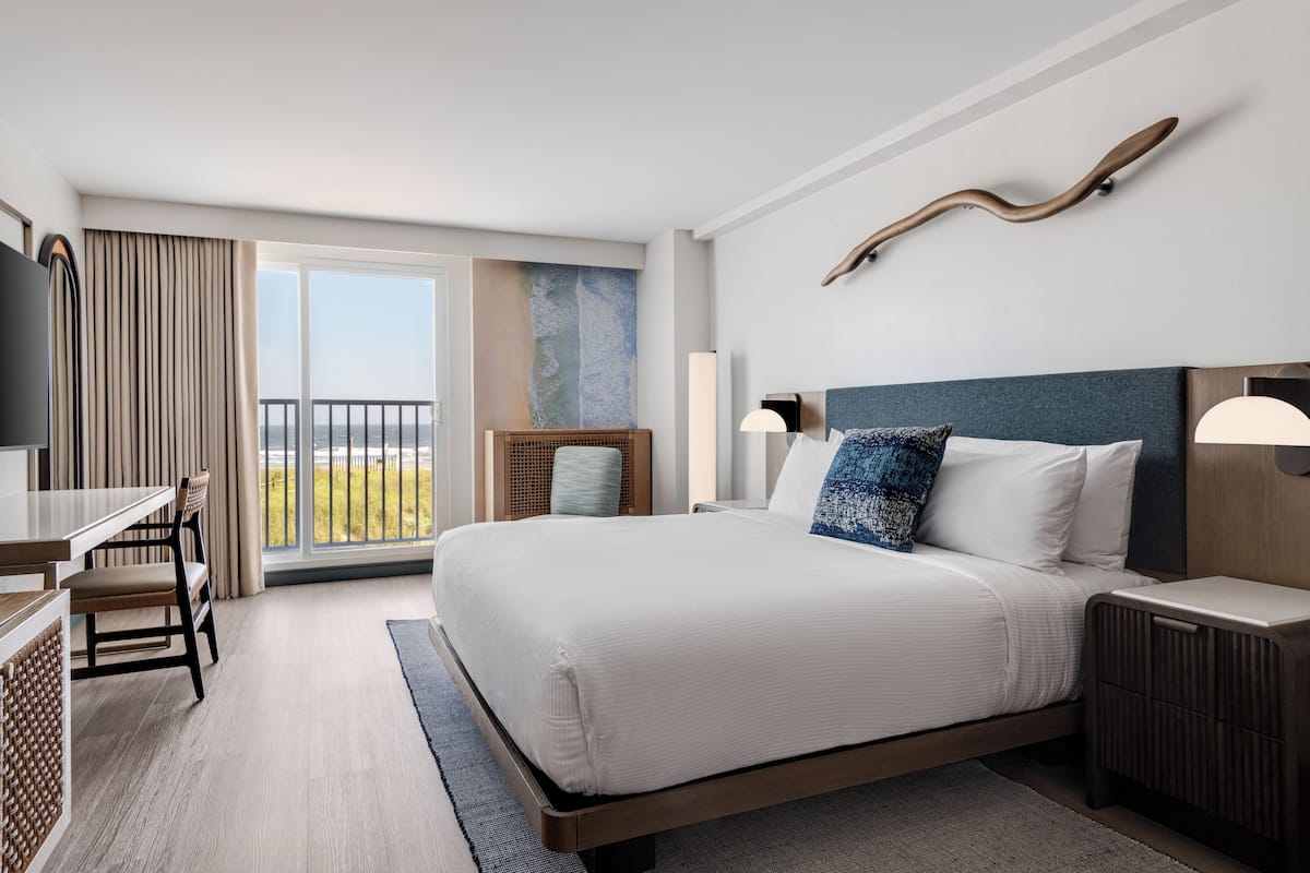 A hotel room with a large bed, a desk, and a chair. There is a balcony with a view of the ocean.