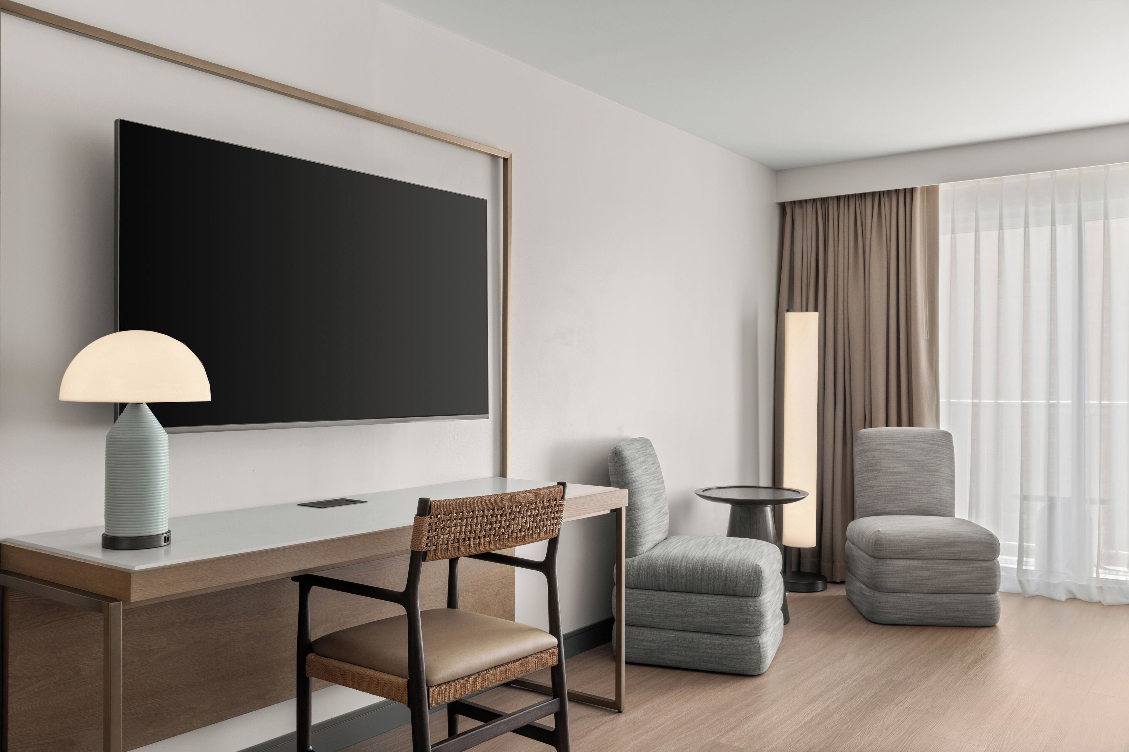 Ein modernes Hotelzimmer mit einem großen Flachbildfernseher, einem Schreibtisch mit Lampe, zwei Polsterstühlen und einem großen Fenster mit leichtem Vorhang.