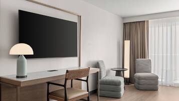 Ein modernes Hotelzimmer mit einem großen Flachbildfernseher, einem Schreibtisch mit Lampe, zwei Polsterstühlen und einem großen Fenster mit leichtem Vorhang.