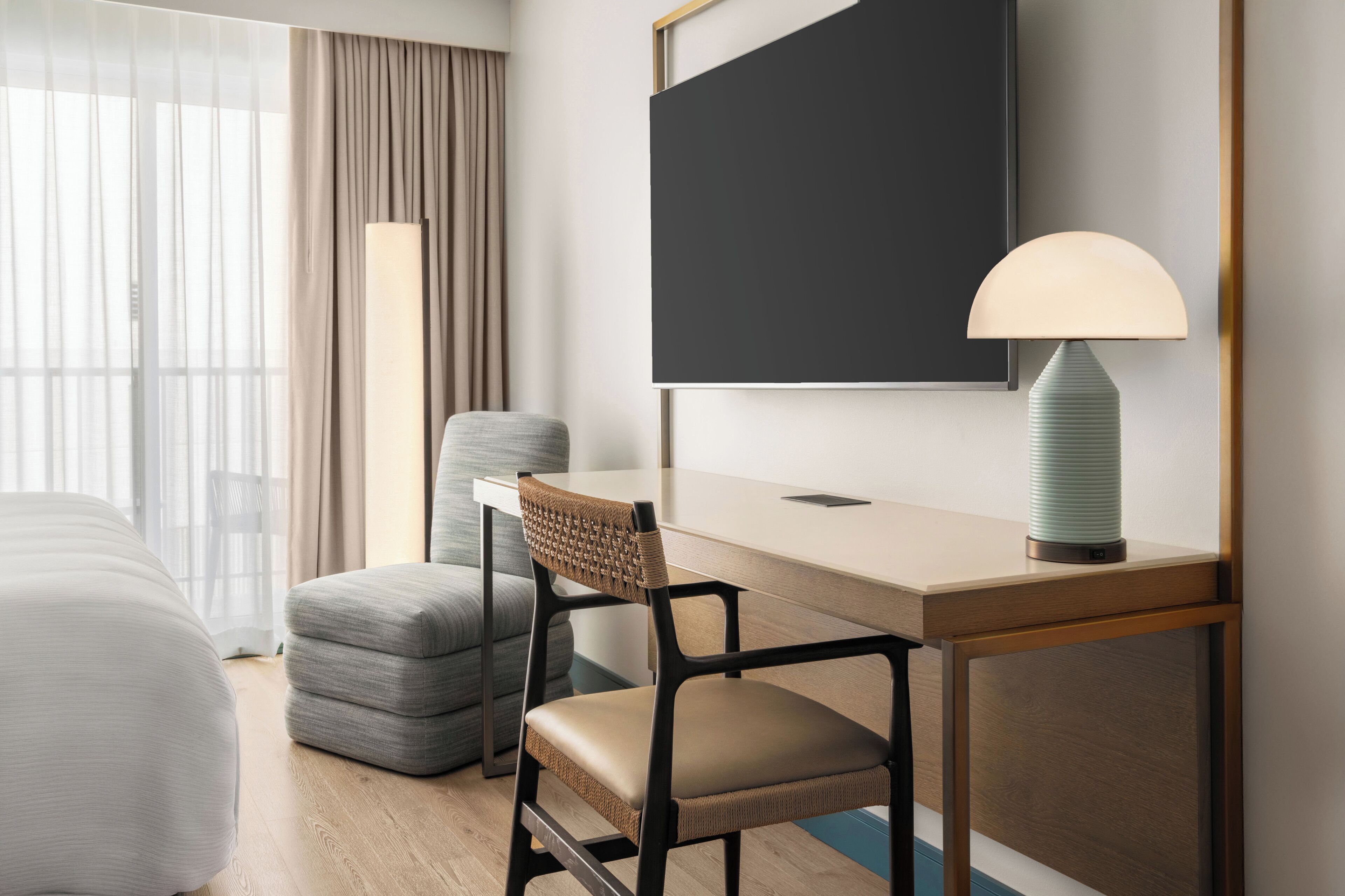 A modern hotel room with a bed, a desk with a lamp, a chair, and a large window with curtains.