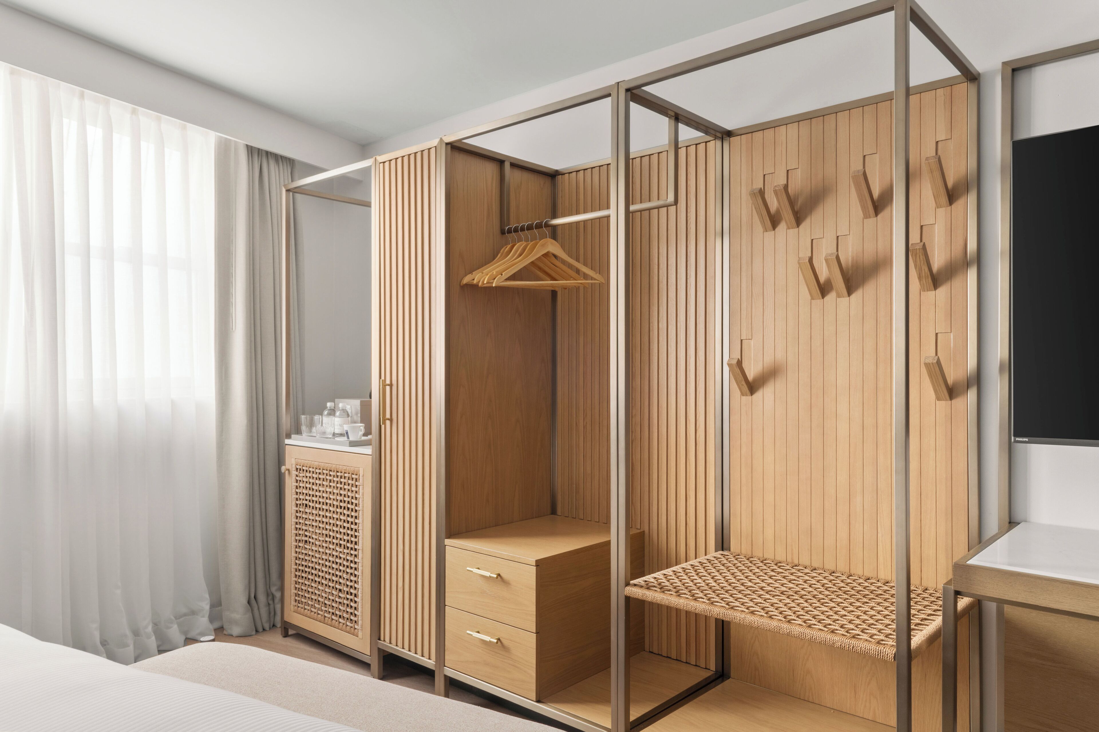 A modern bedroom with a wooden wardrobe, a bedside table, and a window with sheer curtains.