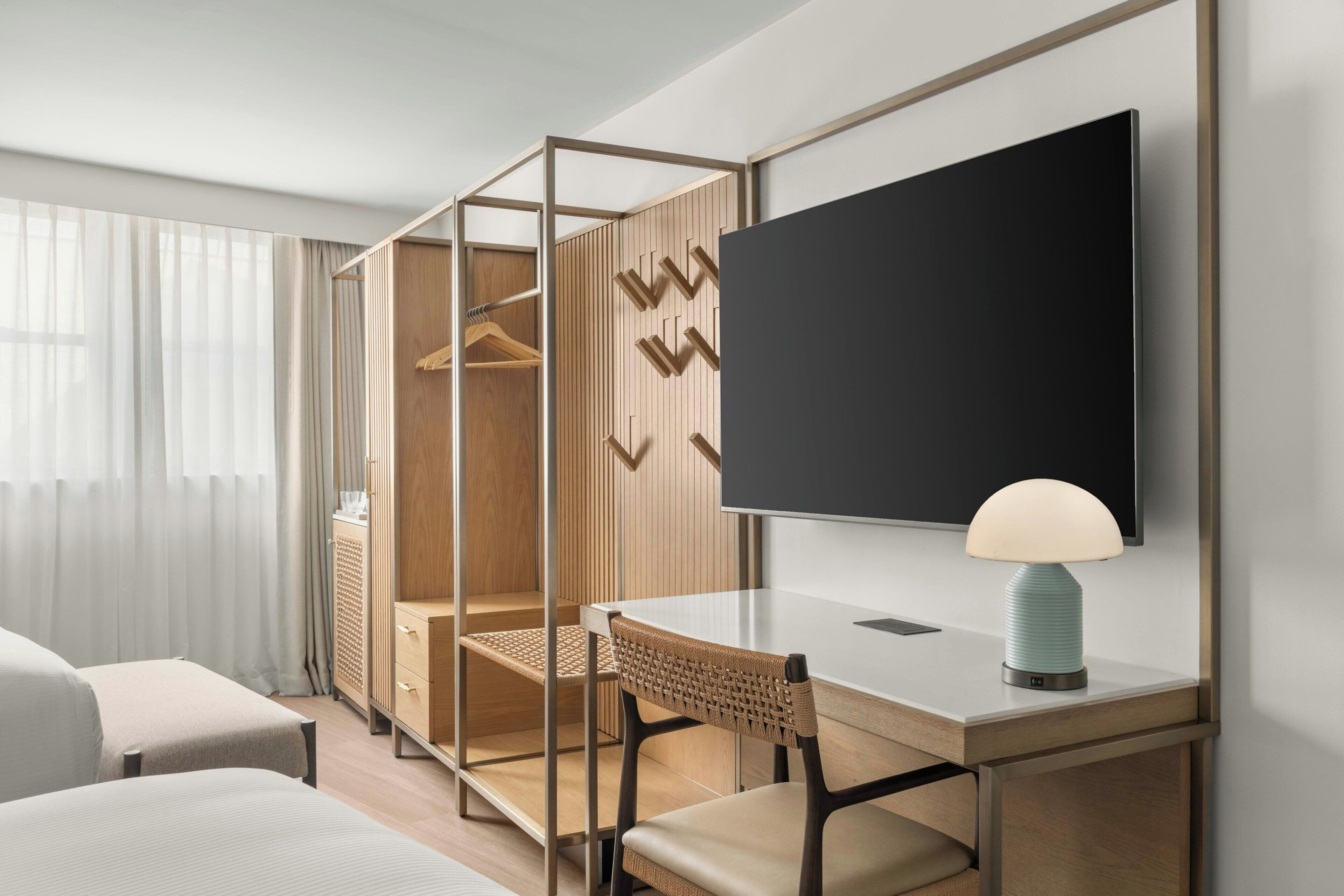 Quarto de hotel moderno com escrivaninha, cadeira, televisão e guarda-roupa com organizador interno.