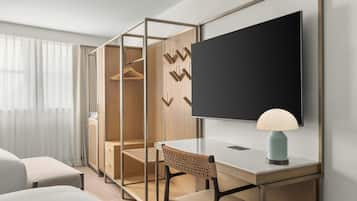 Quarto de hotel moderno com escrivaninha, cadeira, televisão e guarda-roupa com organizador interno.