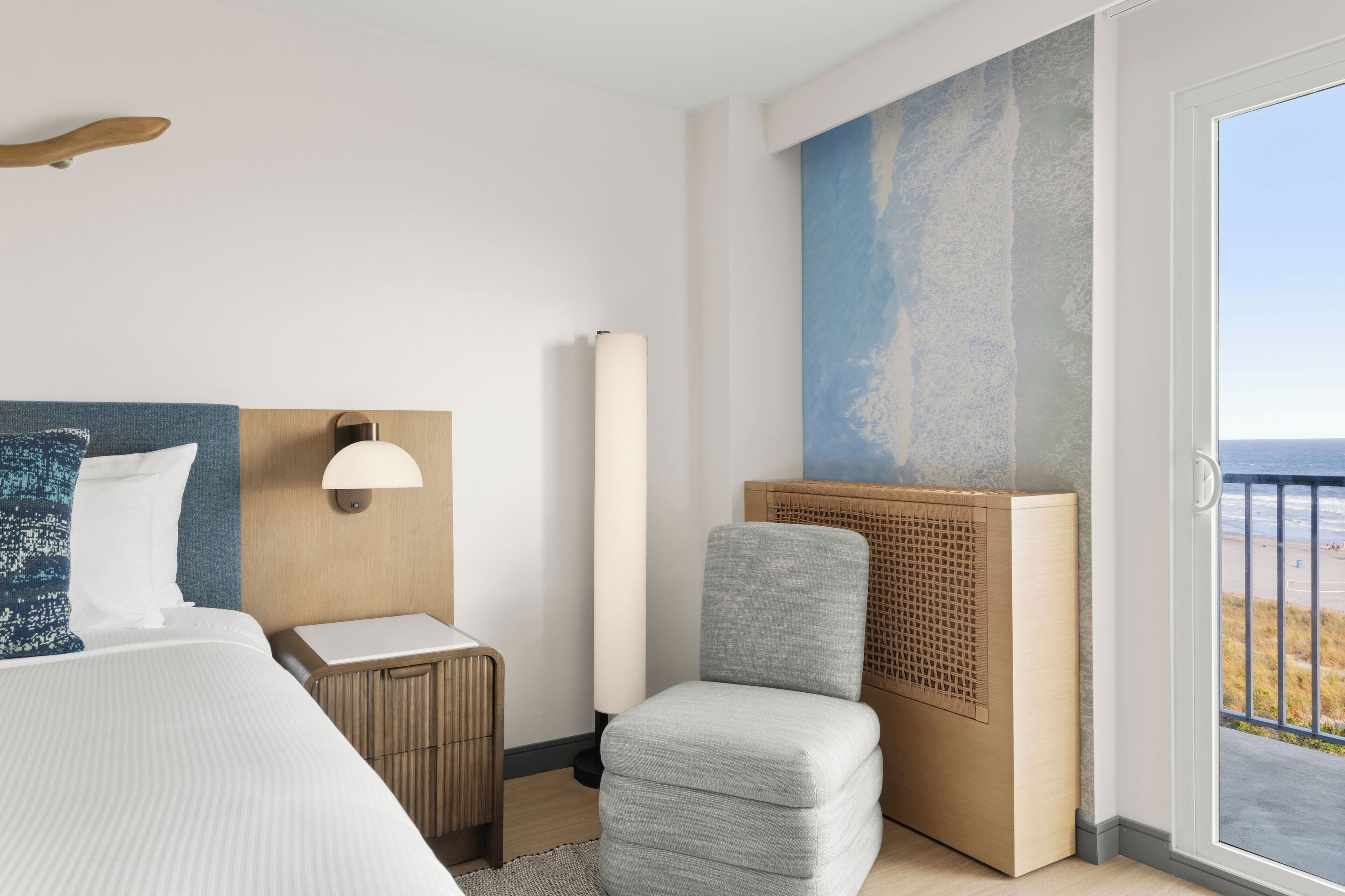 Une chambre d’hôtel avec un lit, une chaise, une table de chevet, une lampe et une grande fenêtre donnant sur la plage.