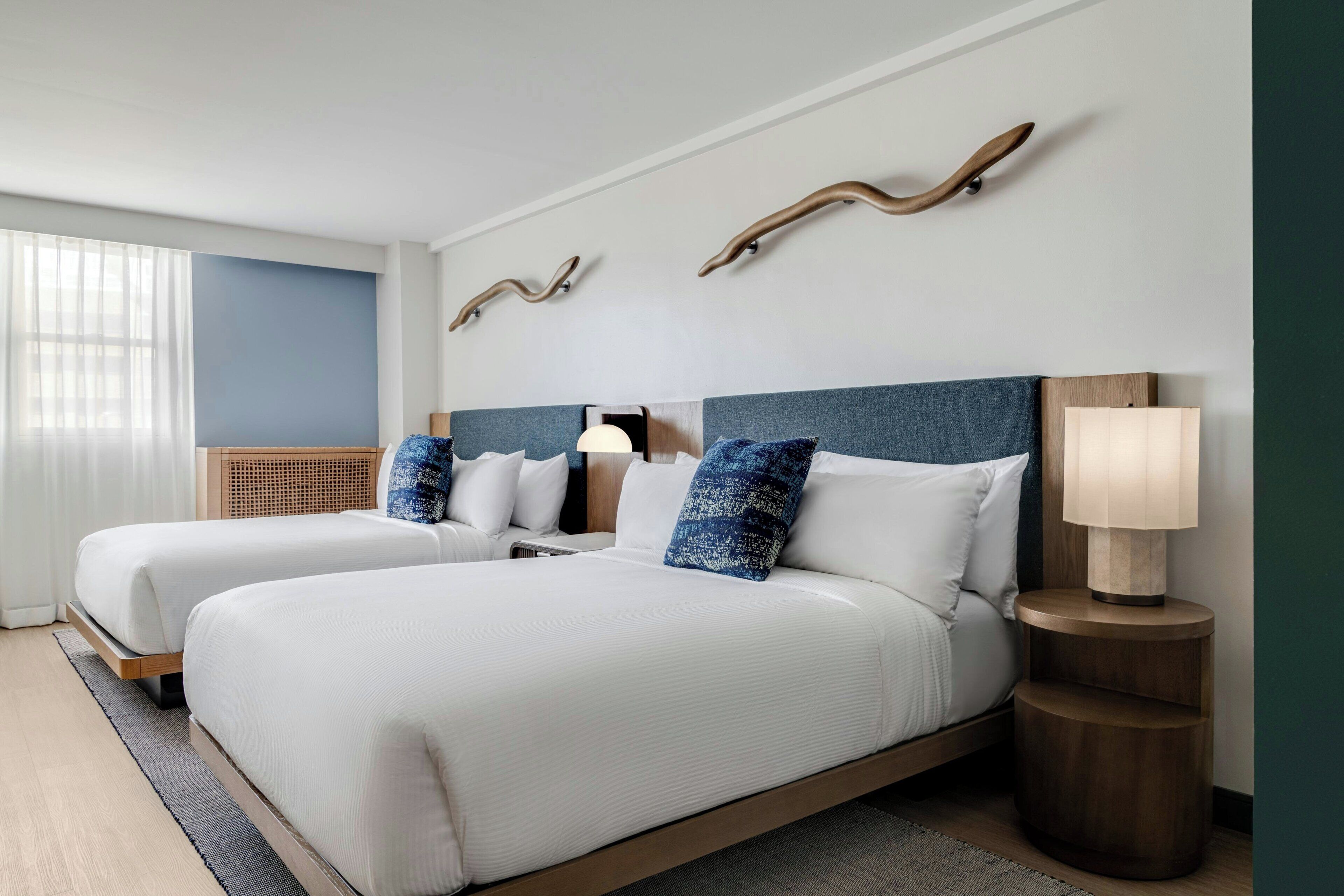 Quarto de hotel com duas camas, uma mesa de cabeceira com abajur e uma janela com cortinas translúcidas.
