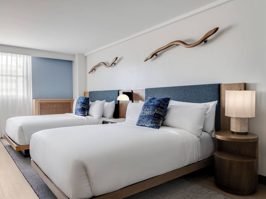 A modern hotel room with two beds, a wooden headboard, a bedside table with a lamp, and a large window with sheer curtains.