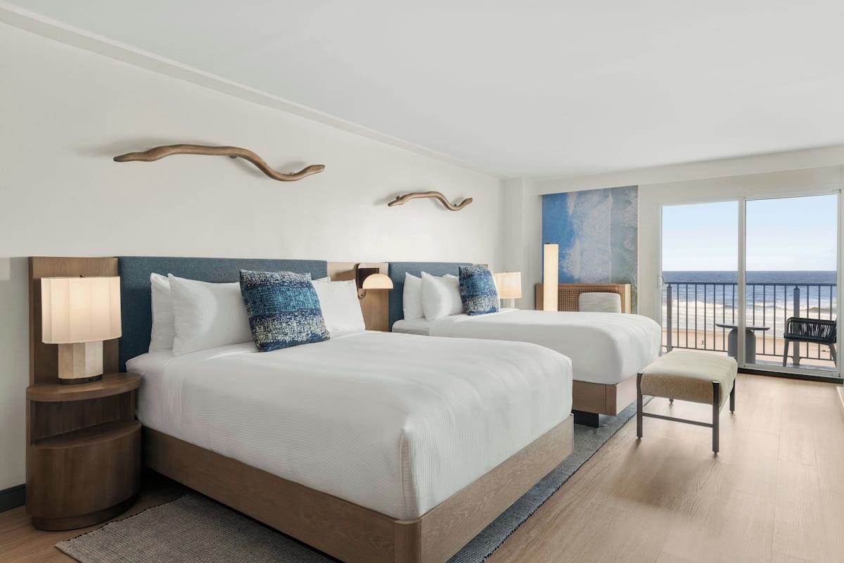 A hotel room with two beds, a balcony with a view of the beach, and a large window.