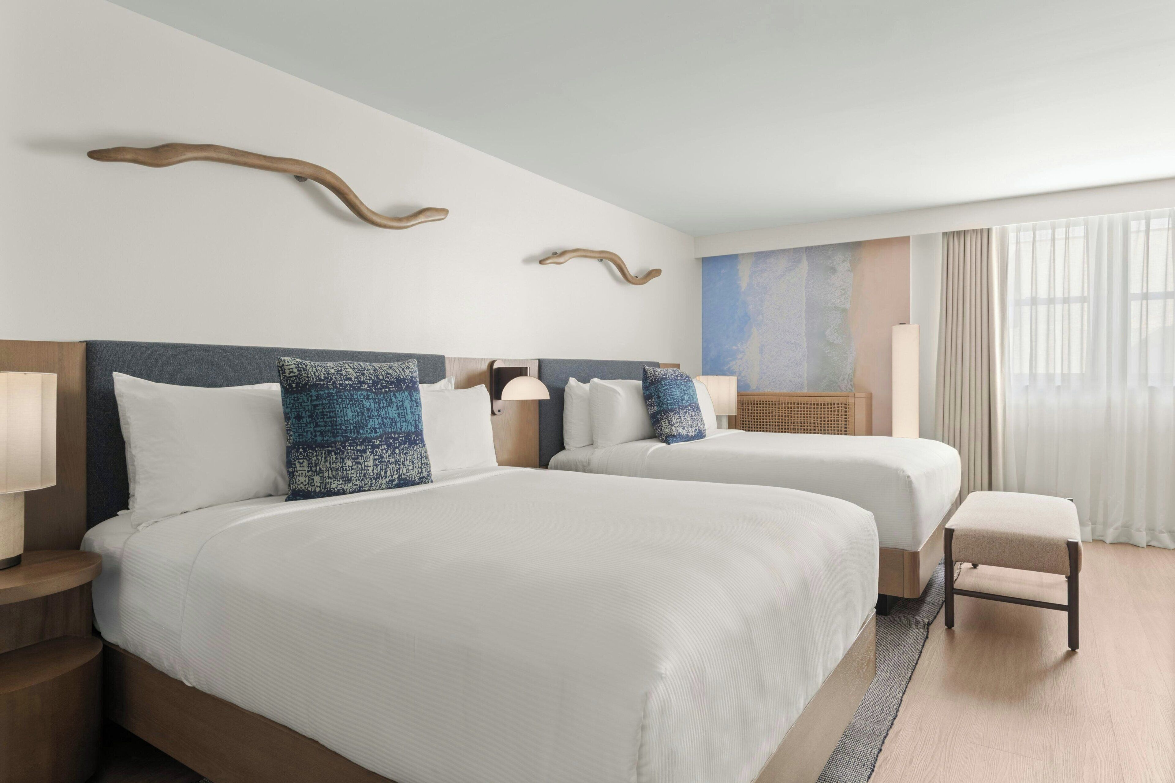 Habitación de hotel con dos camas, cabecero de madera, un pequeño banco y un ventanal con cortinas.