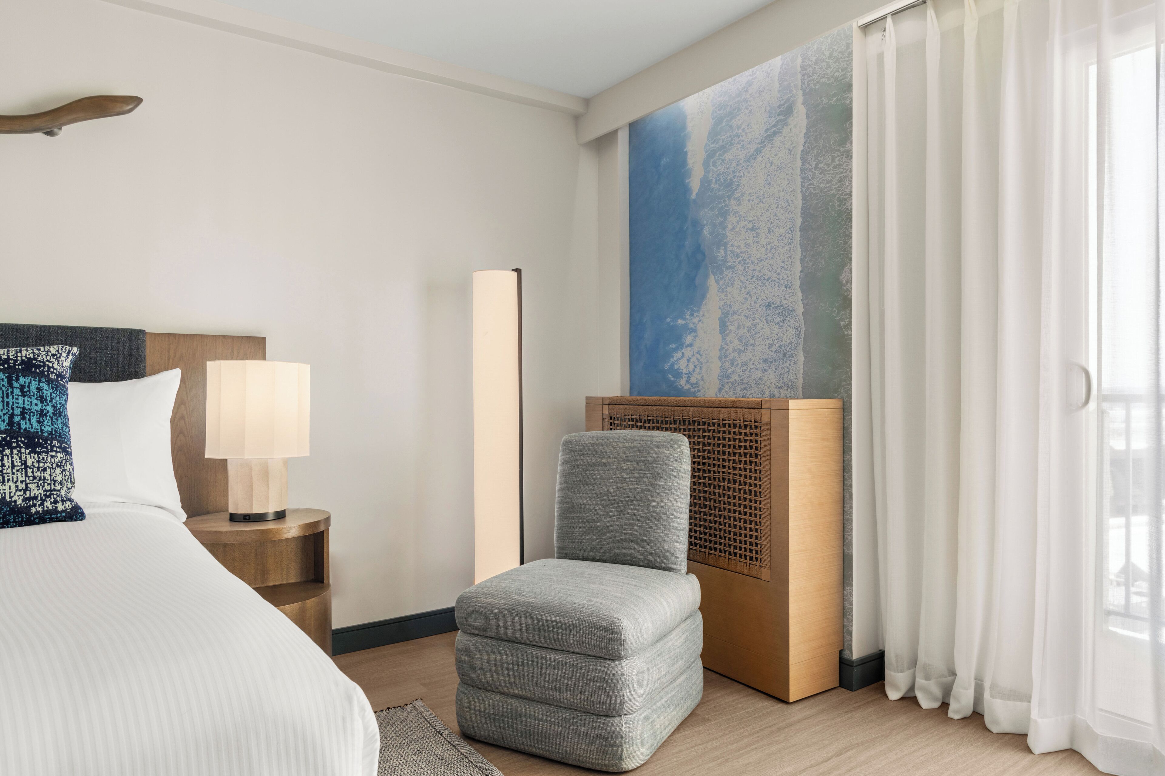 Habitación de hotel con cama, mesita de noche, lámpara, una silla, un cuadro abstracto de gran tamaño y cortinas traslúcidas.