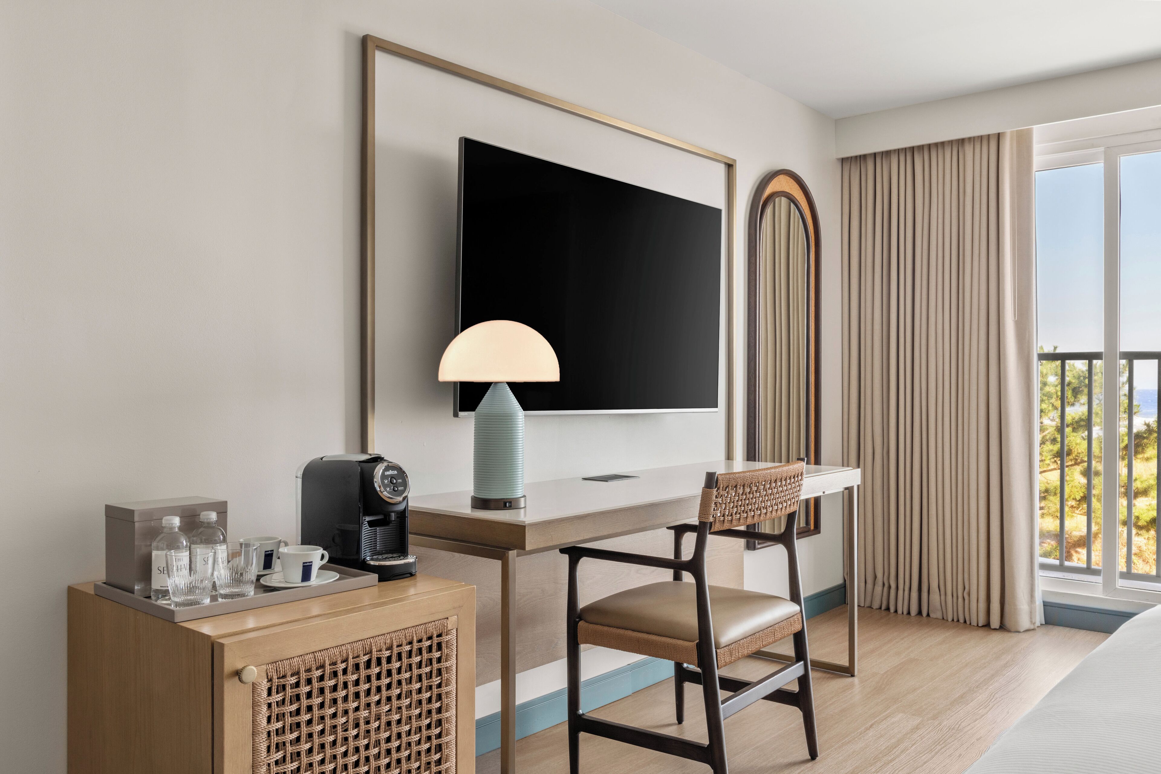 A hotel room with a large flat-screen TV, a desk with a lamp, a chair, a coffee machine, and a minibar.