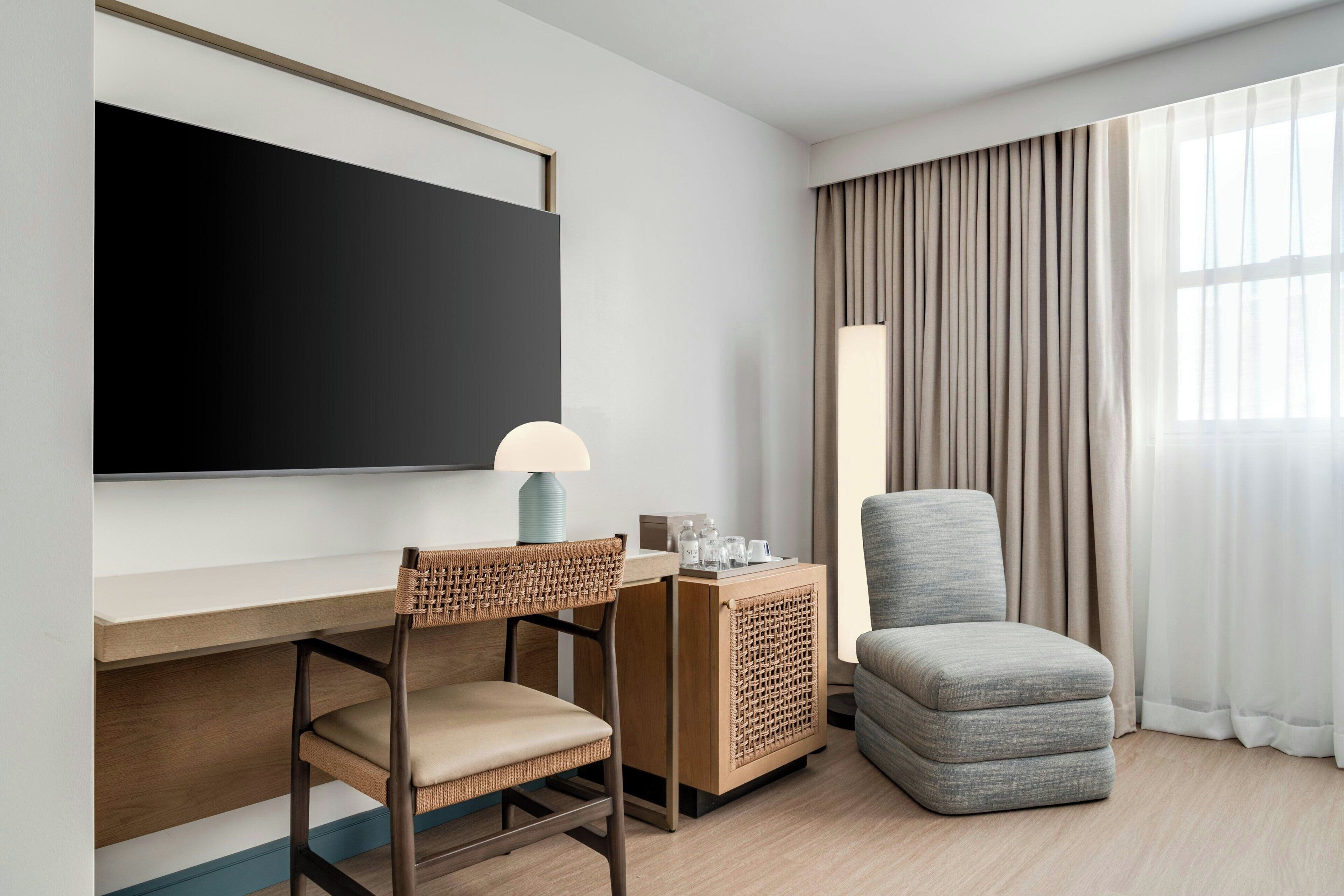 Habitación de hotel con televisor de pantalla plana, escritorio con lámpara, silla de mimbre, sillón mullido y un ventanal con cortinas.