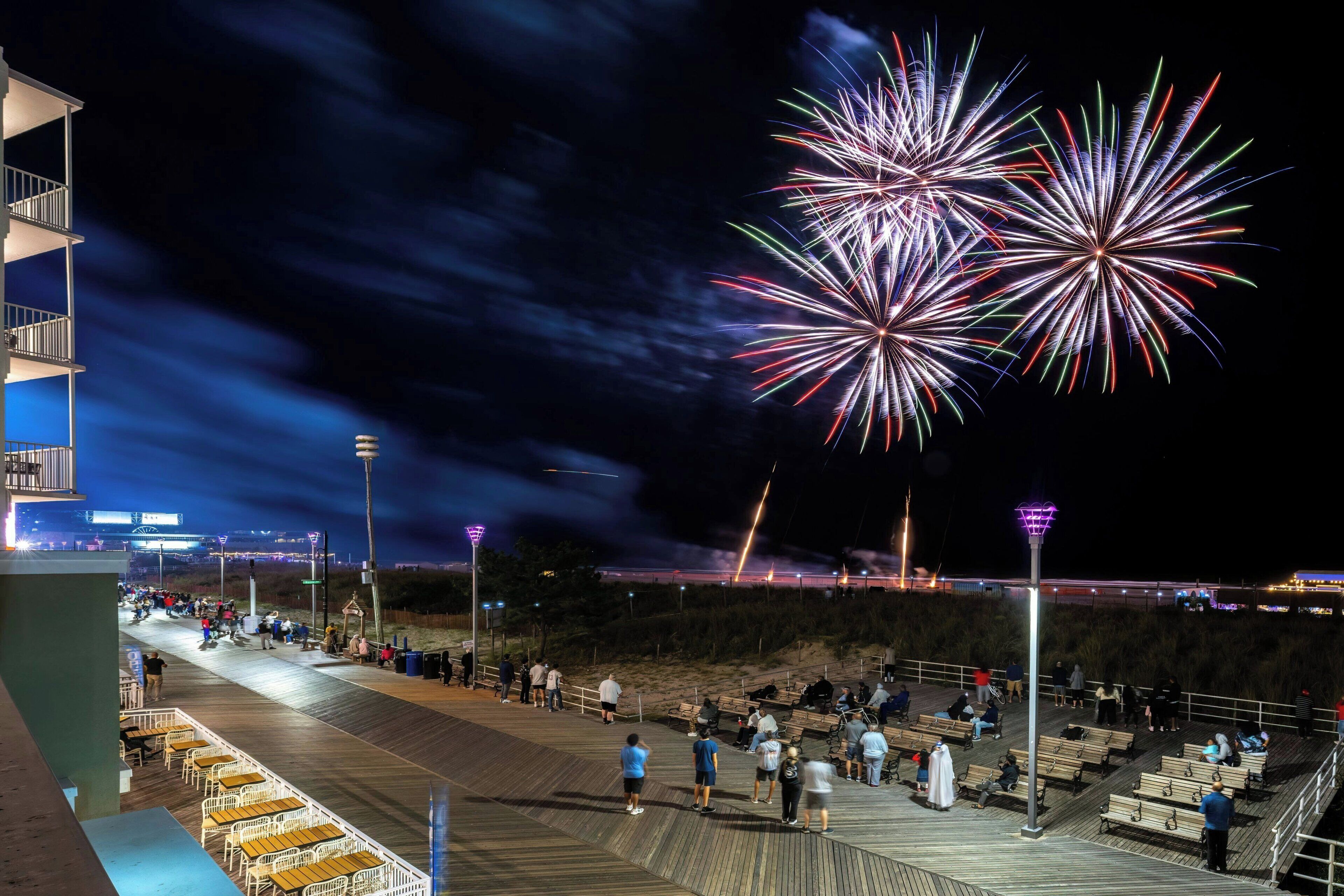 Une promenade en bois bordée de bancs, où l’on peut s’asseoir et observer la foule, avec en toile de fond une plage et des feux d’artifice dans le ciel.