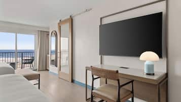 Quarto de hotel com cama, escrivaninha com abajur, cadeira, espelho grande e vista para a praia.