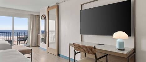 Habitación de hotel con una cama, un escritorio con lámpara, una silla, un espejo grande y vista a la playa.