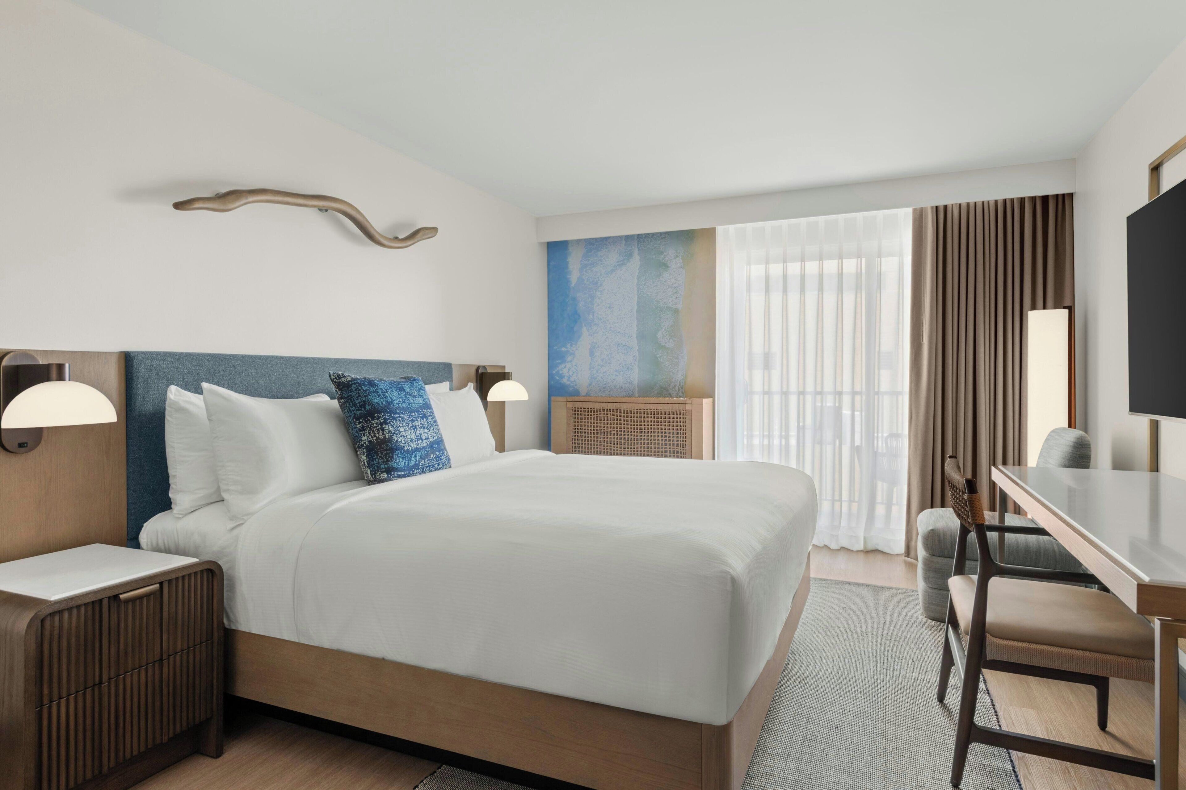 A hotel room with a large bed, bedside tables, a desk with chairs, a flat-screen TV, and a large window with curtains.