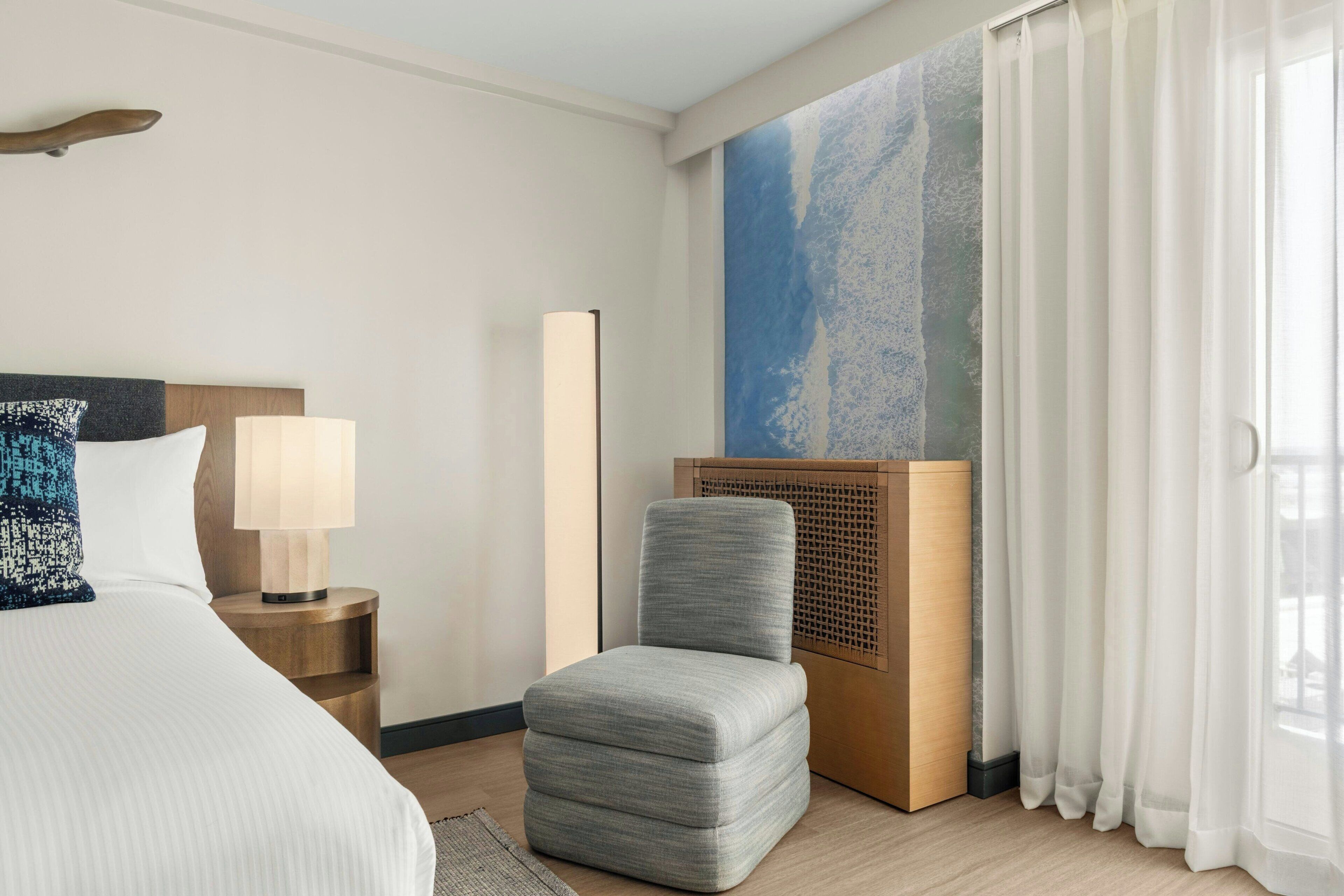 A hotel room with a bed, a side table with a lamp, a grey upholstered chair, a large abstract painting on the wall, and sheer curtains.