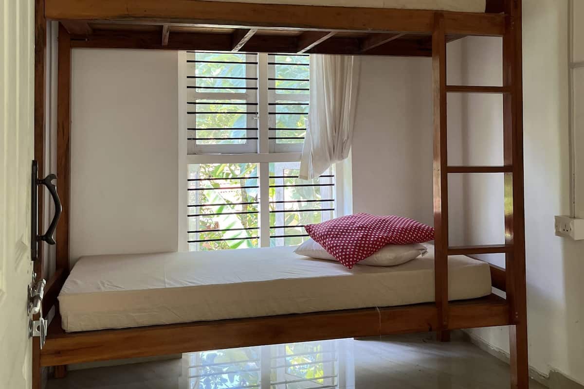 A bunk bed with a wooden frame, a window with blinds, a red polka dot pillow, and a ladder.
