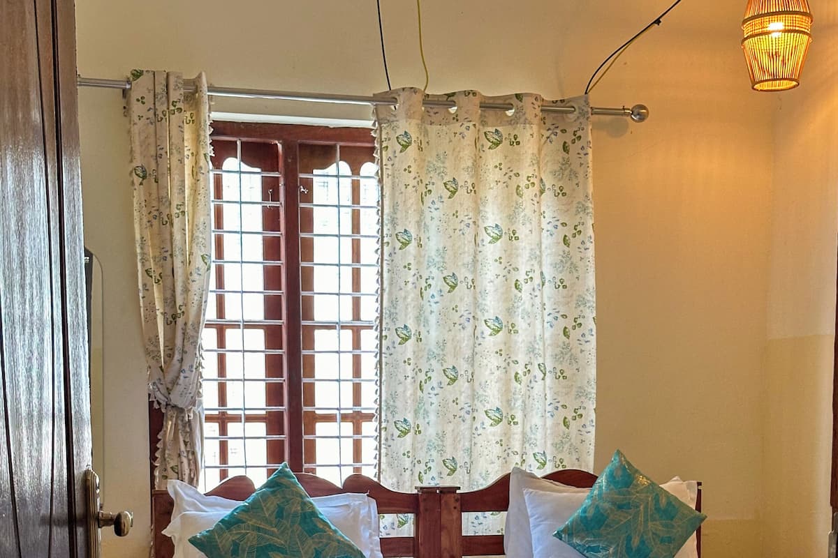 A double bed with a wooden frame, two pillows, a window with patterned curtains, and a hanging light fixture.
