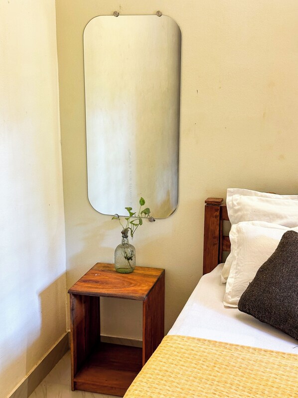 Une chambre à coucher avec un lit, une petite table de chevet en bois sur laquelle est posé un vase de fleurs, et un miroir fixé au mur.