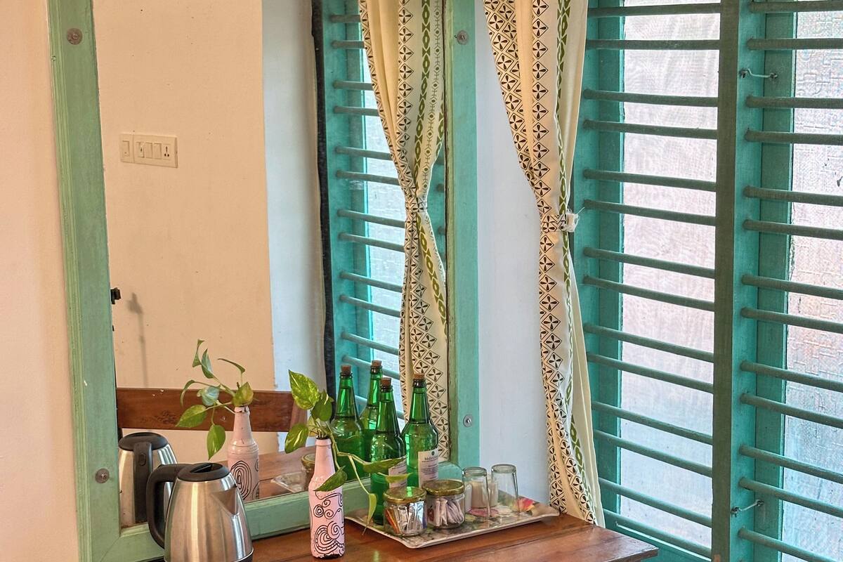 A wooden table with a kettle, a pink vase with a plant, and a tray with bottles and glasses. A mirror with a green frame reflects the room.