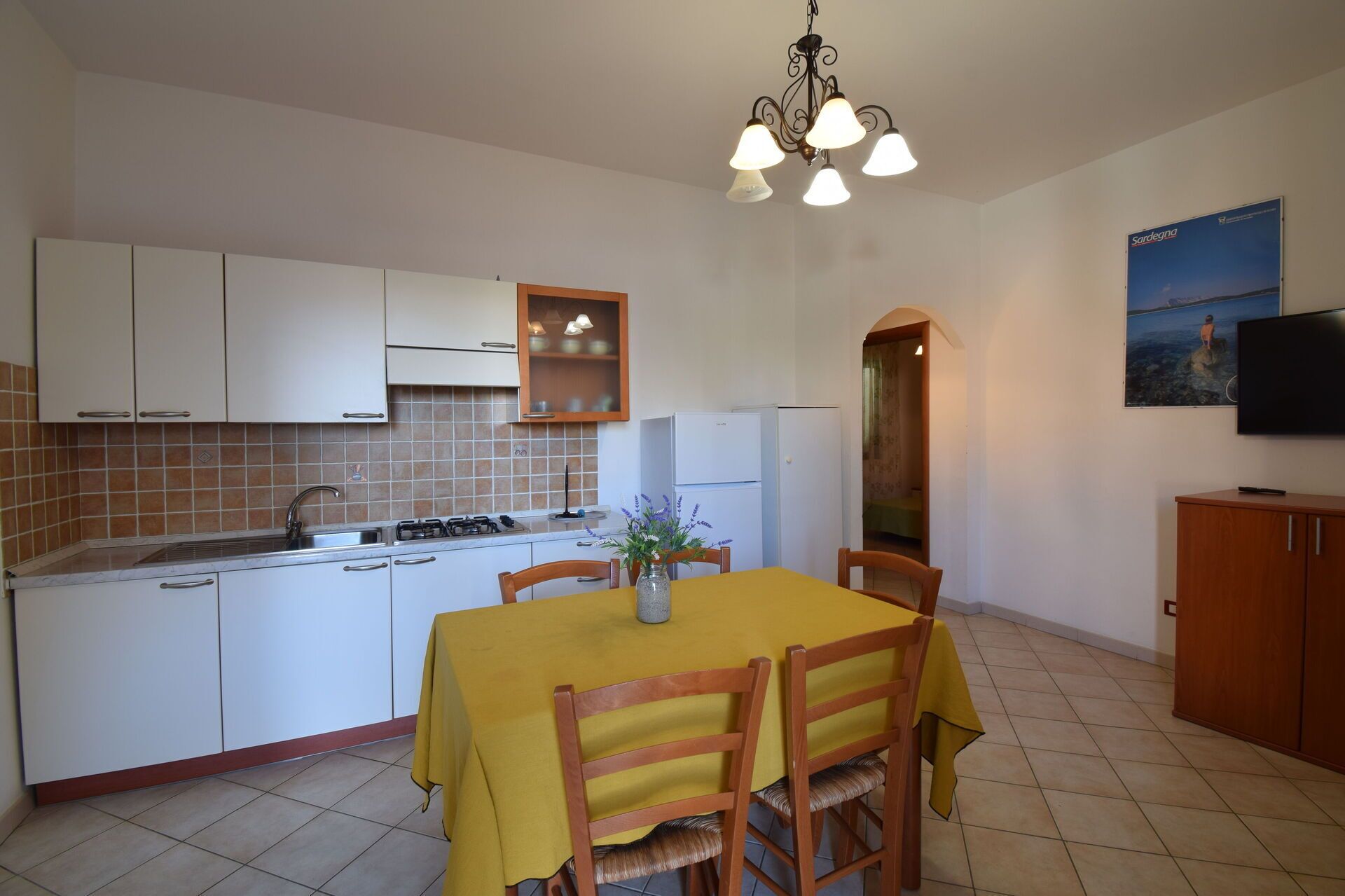 Una cucina compatta con mobili bianchi, un piano cottura a gas e un lavello. C'è una zona pranzo con un tavolo giallo e sedie di legno. Un televisore è montato a parete ed è visibile un quadro.