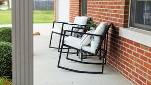 A patio with two white rocking chairs and a small table, set against a brick wall with a window above. - Abe's Hideaway - Arcade, Hot Tub, Home Theater - Quiet upscale neighborhood (Springfield)