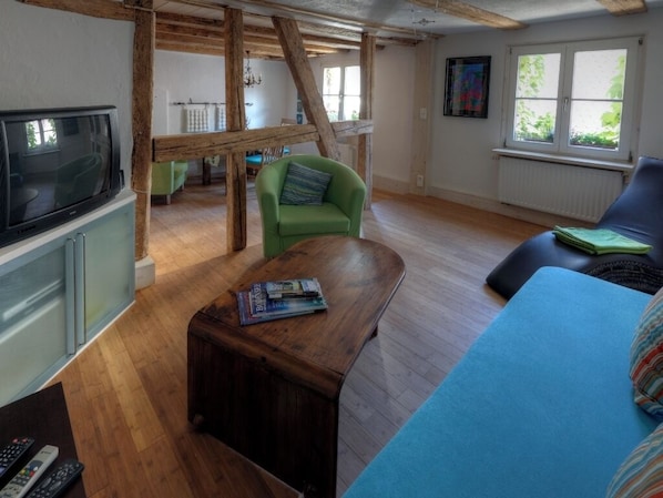 A room with a wooden ceiling, a green armchair, a blue sofa, a wooden coffee table with magazines, and a television on a cabinet.
