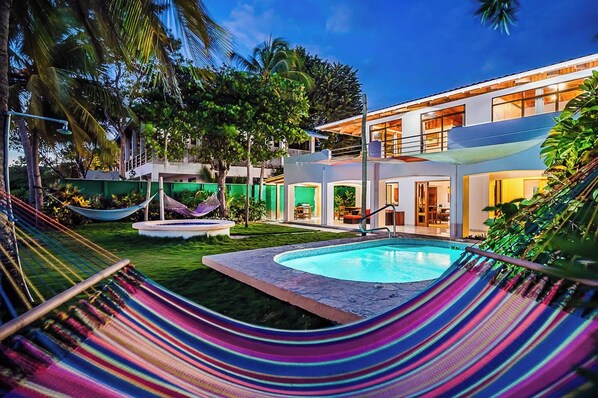 A modern house with a swimming pool, a hammock, and lush greenery.
