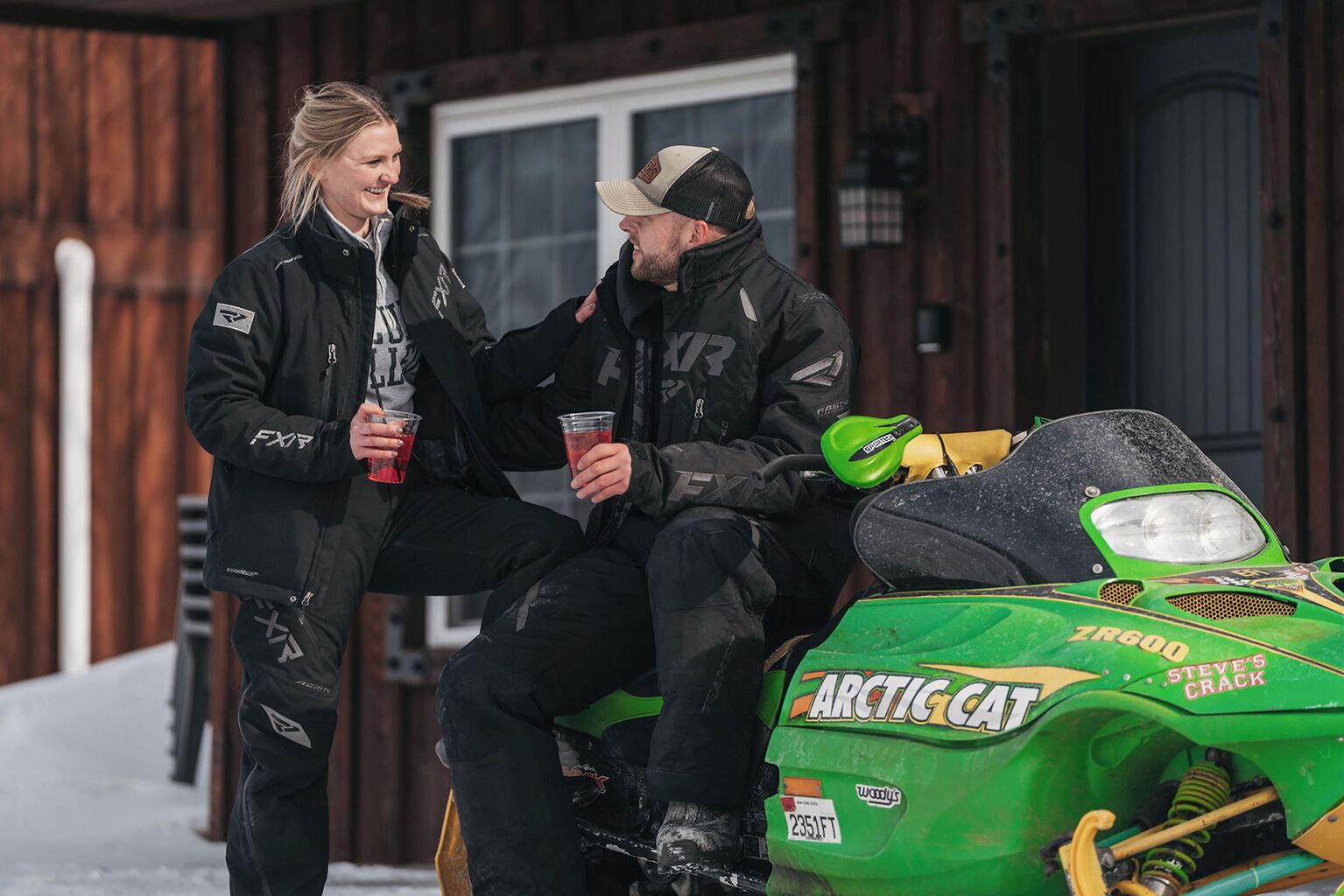 Deux personnes sont assises sur un motoneige vert de la marque « Arctic Cat », devant un bâtiment en bois doté d’une porte et de fenêtres.