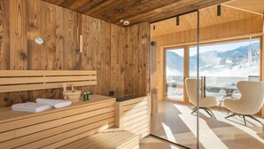 En träinredd interiör med en bastu, en glasdörr som leder ut till en fantastisk utsikt över ett snötäckt berg och moderna sittmöbler.