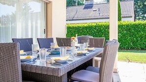 An outdoor dining area with wicker furniture and a glass table, surrounded by a hedge and a building with a tiled roof.