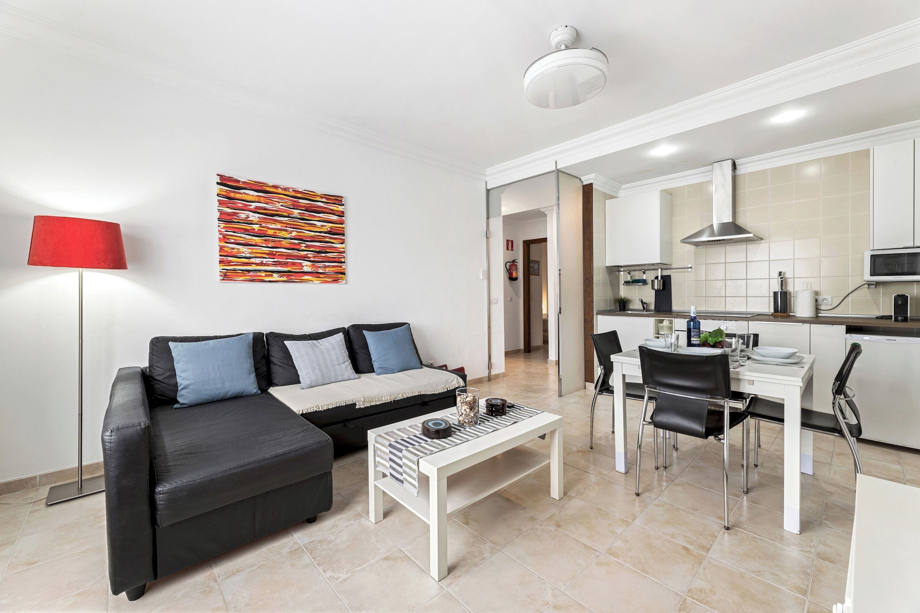 Een moderne woonkamer met een zwarte bank, een witte salontafel en een rode vloerlamp. In de achtergrond is een keuken te zien met witte kasten en een magnetron.