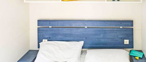 A bed with white bedding, a blue headboard, and a shelf above with a yellow pillow and a pair of sunglasses.