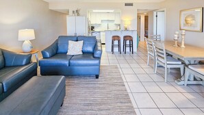A modern living room with a blue sofa, a small table with a lamp, and a kitchen in the background. -  Spacious Ormond Oceanfront  3 bedroom Condo (Ormond Beach)