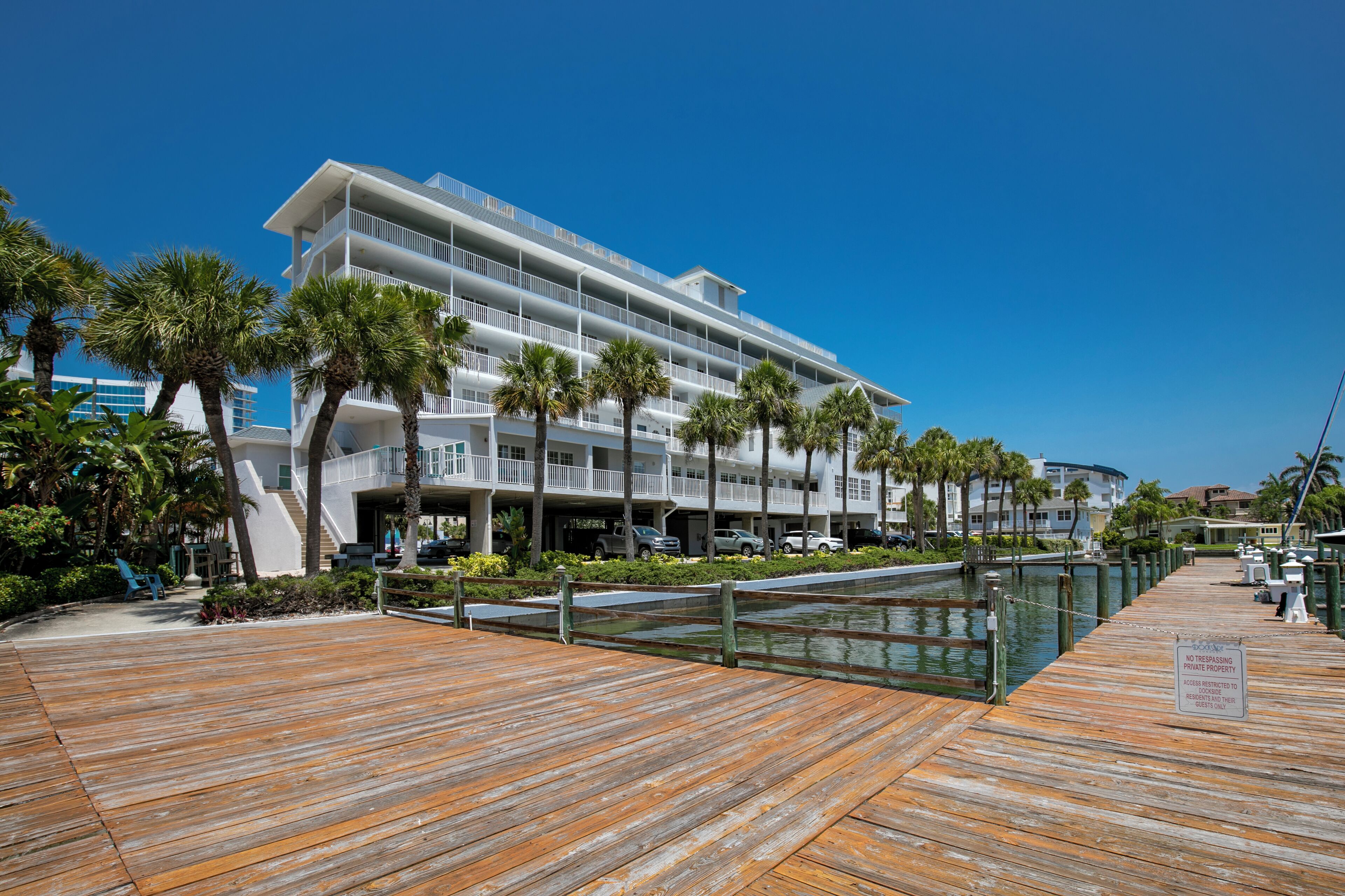 A waterfront hotel with a wooden deck, palm trees, and a clear blue sky.