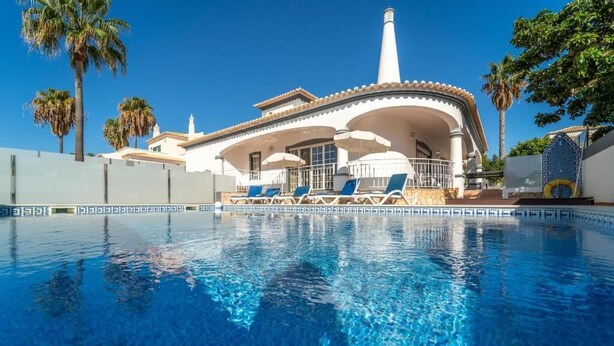Vista de una piscina frente a una casa con fachada blanca, una chimenea cónica y un cielo azul claro.