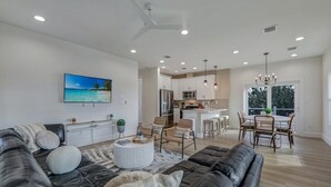 A modern living room with a black leather sofa, a dining area with a round table and chairs, a kitchen with white cabinets, and a large window providing a view of the outdoors. - Shell Shack - New Luxurious Home (Sanibel)