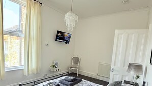 A bedroom with a bed, a TV, a chandelier, a chair, and a window with curtains.