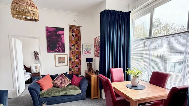 A modern living room with a sofa, a dining table with chairs, a hanging light fixture, and a large window with curtains.