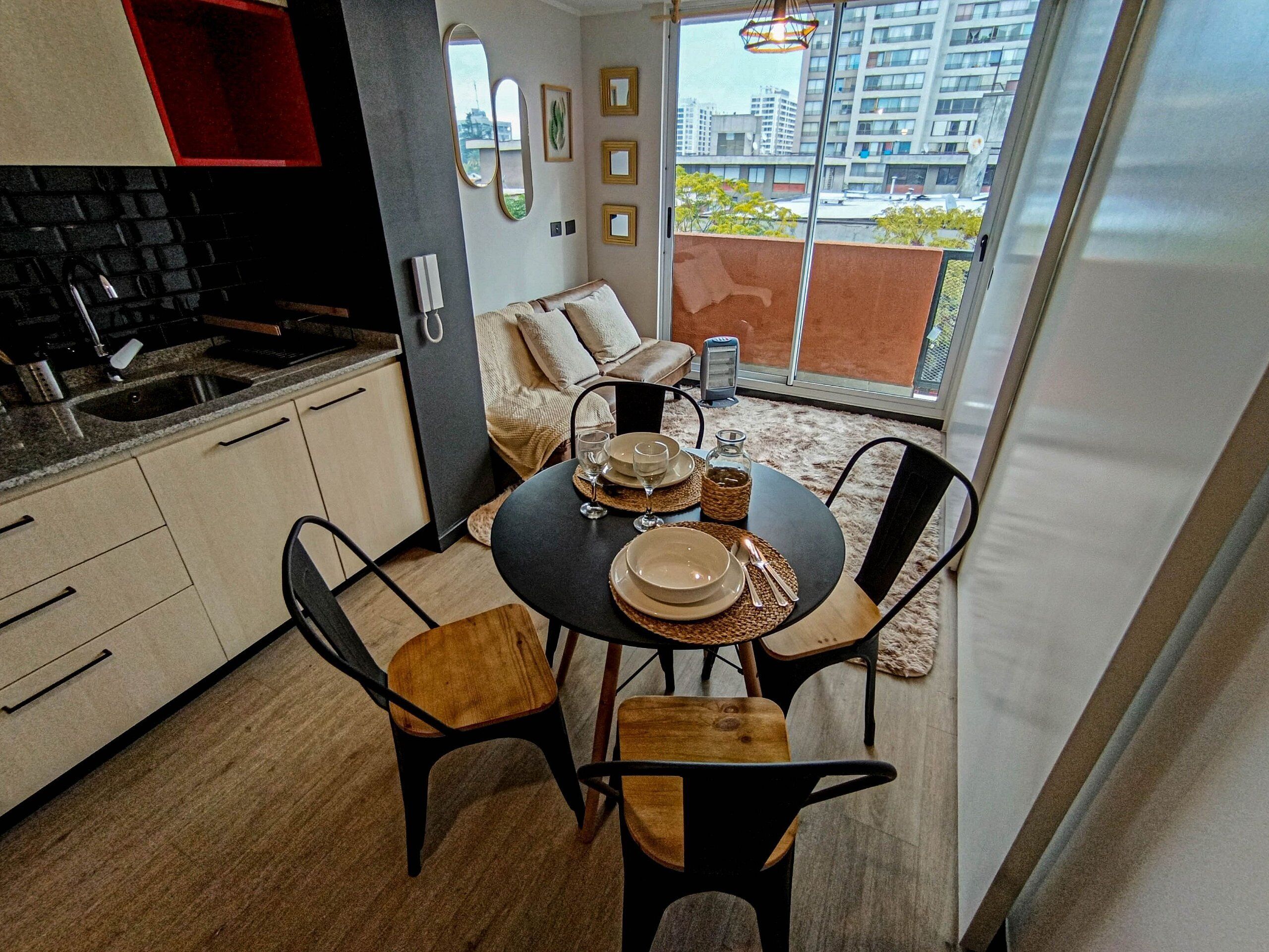 A modern kitchen with a dining area, featuring a round table set for four, a built-in oven, and a view of the city through large windows.