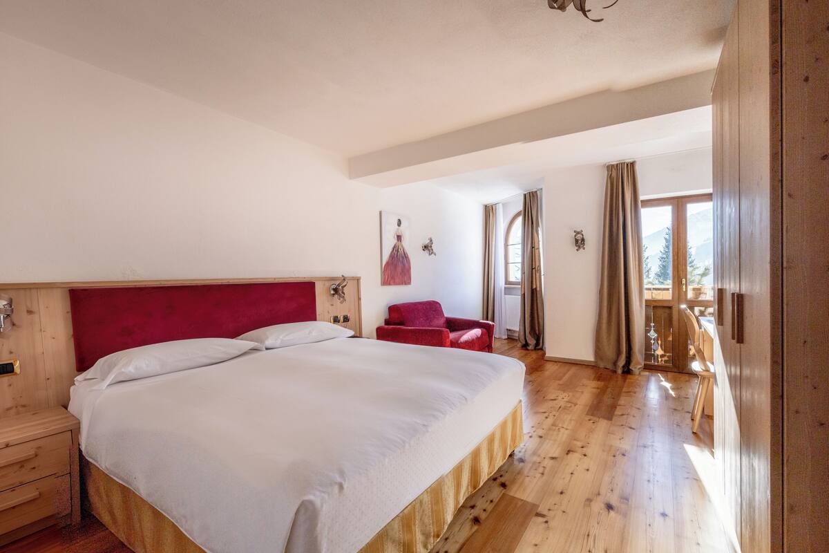 A bedroom with a large bed, wooden headboard, a red chair, and a view of a balcony with curtains.