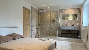 A bedroom with a large bed, a shower with a stone wall, and a bathroom with a sink and mirror. - House to rent for summer holiday weekends for 8 -9-10- 11 - 12 - 13 persons (Compolibat)