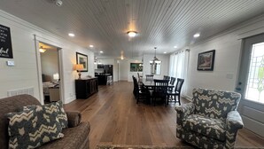 Un salón espacioso con piso de madera, una zona de comedor con una mesa grande y sillas, y una cocina al fondo.
