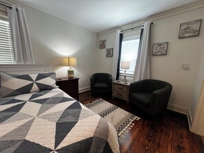 Un dormitorio con cama, dos sillones, una mesita de noche con lámpara, una ventana con cortinas y decoración en la pared.