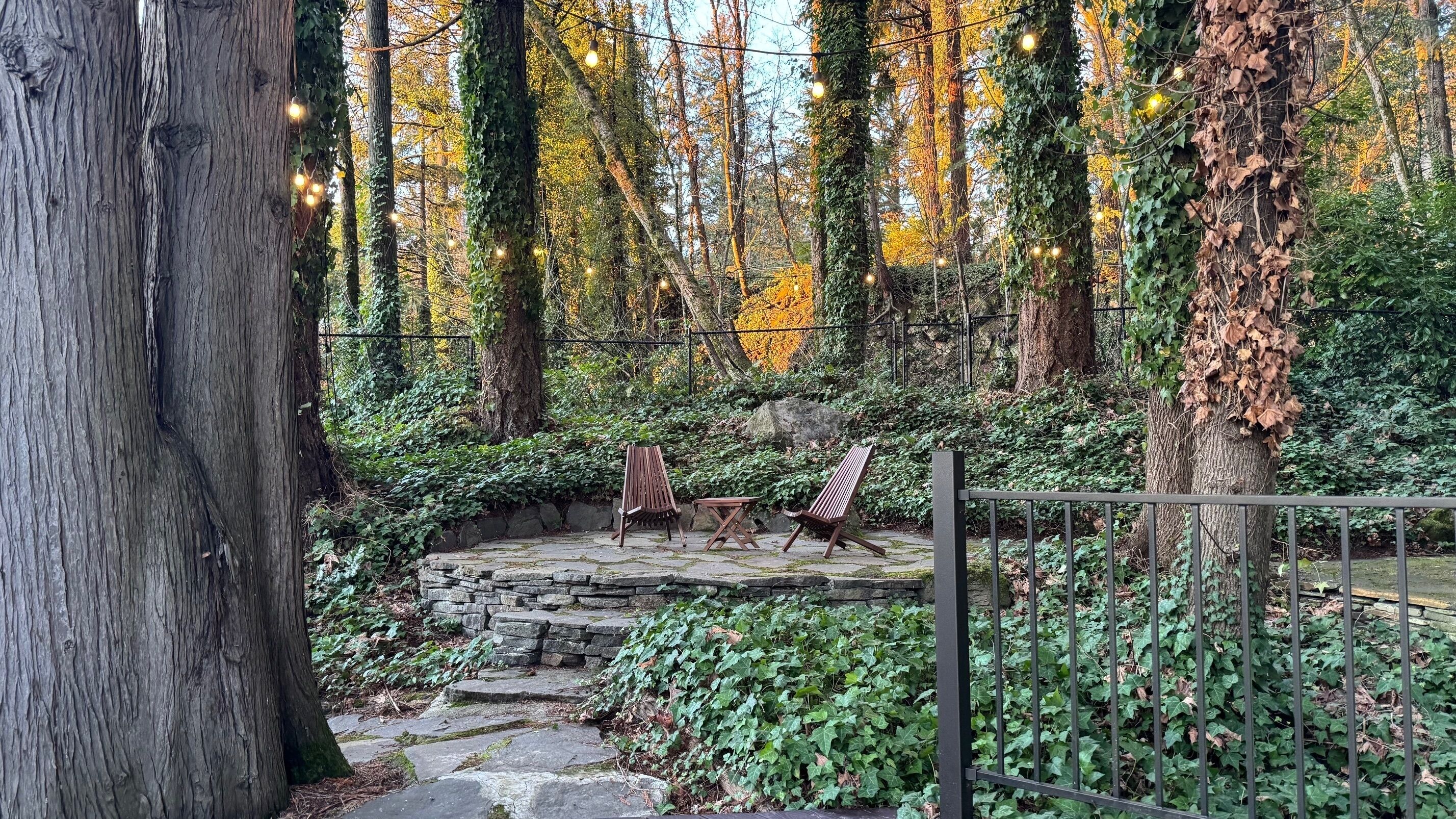 A stone pathway leading to a seating area with three wooden chairs and a table, surrounded by trees and greenery.