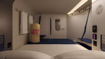 Ein kompakter, moderner Raum mit einem Bett, einem Kopfkissen mit Schlafmodus-Symbol, einem kleinen Fenster und einer Bedienkonsole.